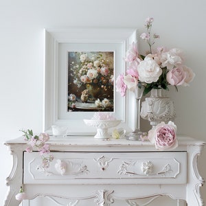 May include: A white vintage-style dresser with a single drawer, featuring ornate carvings. A framed painting of pink roses in a vase sits on top of the dresser, along with a white vase with pink roses and a clear glass vase with pink flowers.