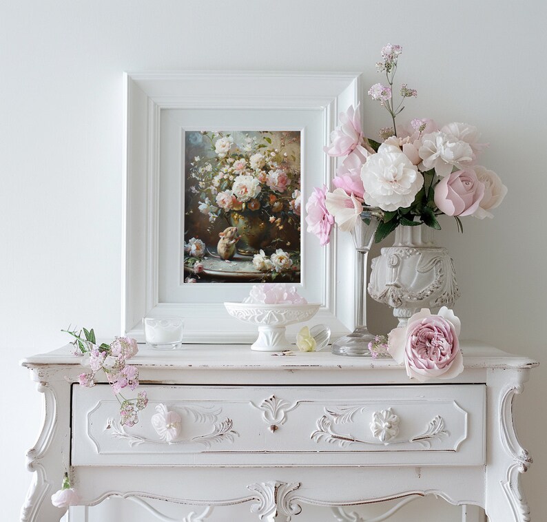 May include: A white vintage-style dresser with a single drawer, featuring ornate carvings. A framed painting of pink roses in a vase sits on top of the dresser, along with a white vase with pink roses and a clear glass vase with pink flowers.
