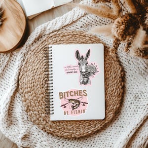 May include: Two stickers on a white notebook. The top sticker is a black and white illustration of a donkey with the text "a little sweet but mostly a smart-ass". The bottom sticker is pink with the text "Bitches be fishin'" and a black and white illustration of a fish.