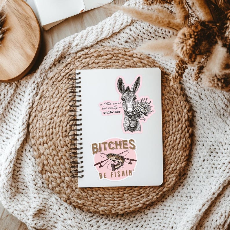 May include: Two stickers on a white notebook. The top sticker is a black and white illustration of a donkey with the text "a little sweet but mostly a smart-ass". The bottom sticker is pink with the text "Bitches be fishin'" and a black and white illustration of a fish.