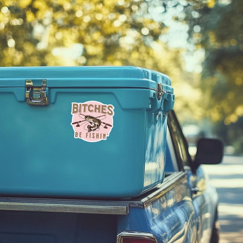 May include: A blue cooler with a pink and gold sticker that says "Bitches Be Fishin'" with a cartoon fish and fishing rods. The cooler is sitting in the bed of a blue pickup truck.