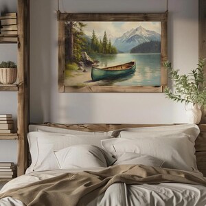 May include: A framed landscape painting depicts a serene lake scene with a green canoe resting on the shore. The artwork features a mountain range in the background, lush green trees, and a clear blue sky. The frame is made of natural wood.