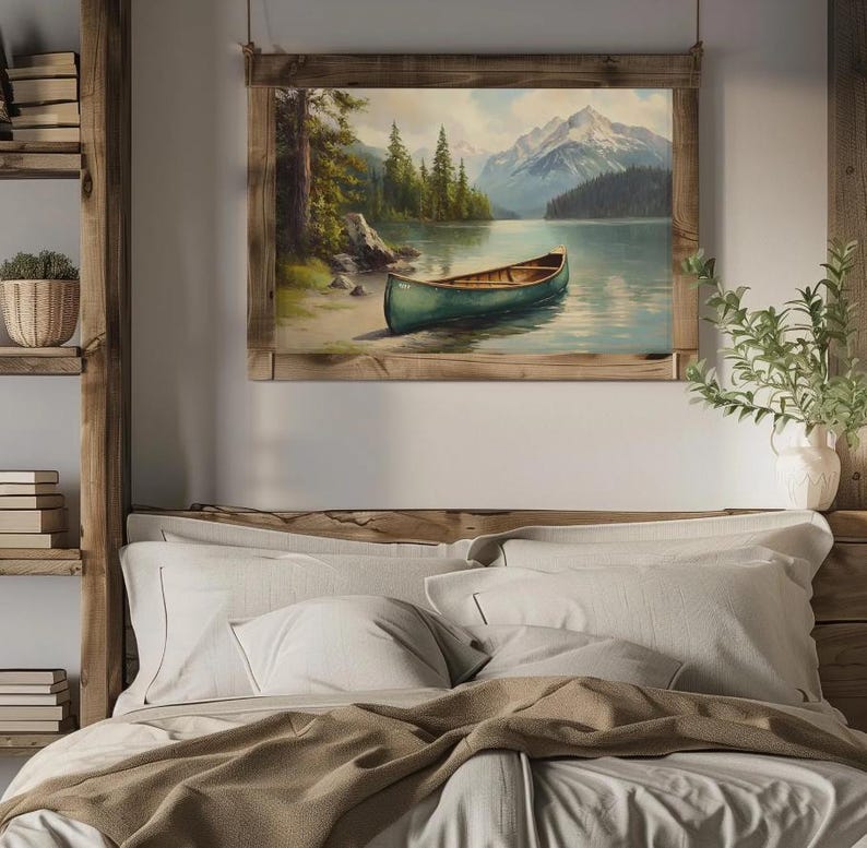 May include: A framed landscape painting depicts a serene lake scene with a green canoe resting on the shore. The artwork features a mountain range in the background, lush green trees, and a clear blue sky. The frame is made of natural wood.