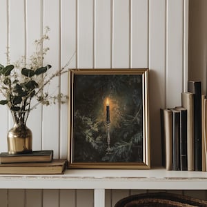 May include: A framed print of a candle burning in a dark forest setting. The candle is on a candlestick and is surrounded by pine branches. The print is in a gold frame and is hanging on a white wall.