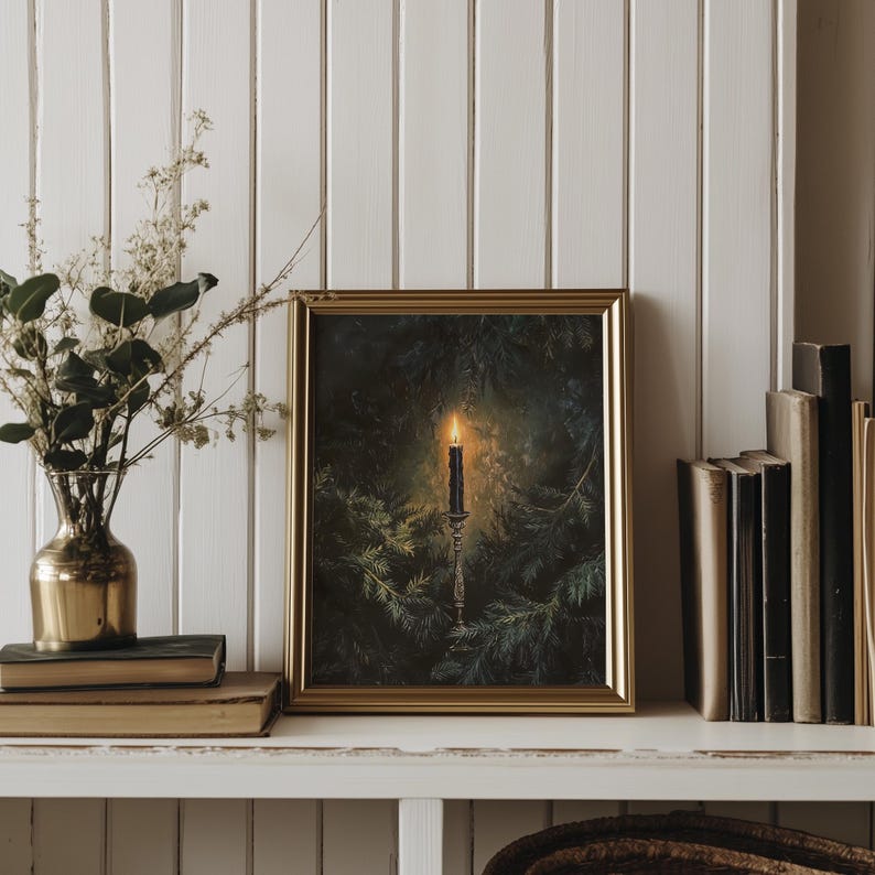 May include: A framed print of a candle burning in a dark forest setting. The candle is on a candlestick and is surrounded by pine branches. The print is in a gold frame and is hanging on a white wall.