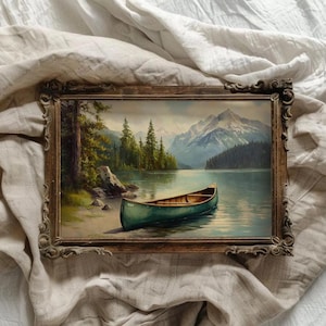 May include: A framed landscape painting depicts a serene lake scene with a green canoe resting on the shore. The artwork features a mountain range in the background, lush green trees, and a wooden frame.