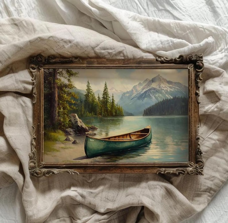 May include: A framed landscape painting depicts a serene lake scene with a green canoe resting on the shore. The artwork features a mountain range in the background, lush green trees, and a wooden frame.