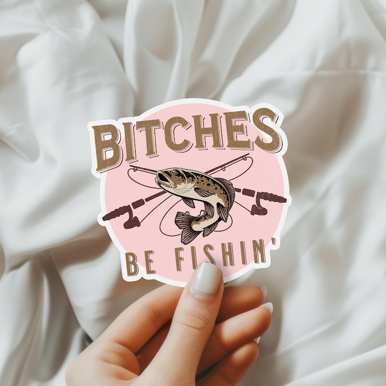 May include: A pink and brown sticker with a fish and fishing rods. The text on the sticker reads "Bitches Be Fishin'"
