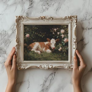 May include: A framed painting featuring a brown and white calf resting in a grassy area surrounded by pink roses. The ornate, cream-colored frame has gold accents. The painting is held by two hands against a marble background.