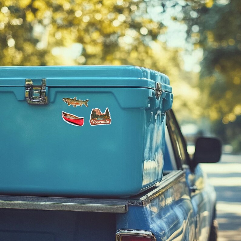 May include: A bright light blue cooler featuring three stickers on its side: a brown fish, a red canoe, and a landscape illustration with the text Yosemite. The cooler has a metallic latch and is positioned on the back of a blue vehicle.