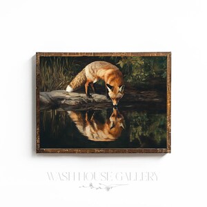 May include: A framed print depicts a fox drinking from a still body of water. The fox has reddish-orange fur and a white-tipped tail. The water reflects the fox and the surrounding foliage, creating a symmetrical image. The frame is a dark brown color.