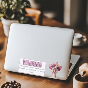 May include: A rectangular sticker on a silver laptop featuring a goose wearing a pink cowboy hat with a white flower and pink glasses. The text reads: WARNING I get distracted by fancy saddles, pretty headstalls, fast, shiny horses, and sparkly things.