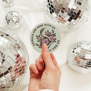 May include: A round sticker with the text "ANIMALS ARE HERE WITH US NOT FOR US" encircling a central design. The design features a snake intertwined with pink roses on a light green background. The sticker is held in front of a background of disco balls.
