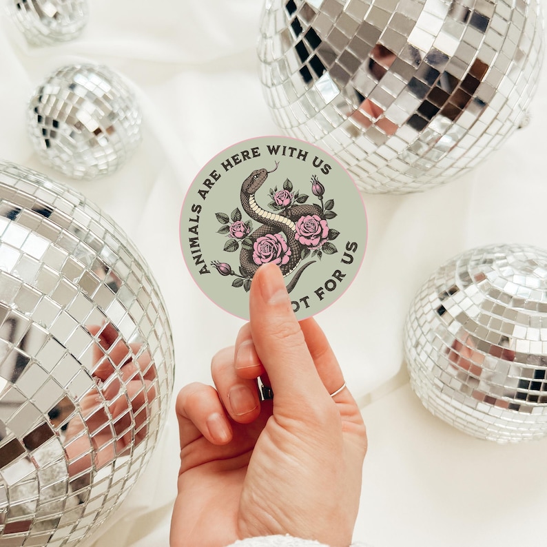 May include: A round sticker with the text "ANIMALS ARE HERE WITH US NOT FOR US" encircling a central design. The design features a snake intertwined with pink roses on a light green background. The sticker is held in front of a background of disco balls.
