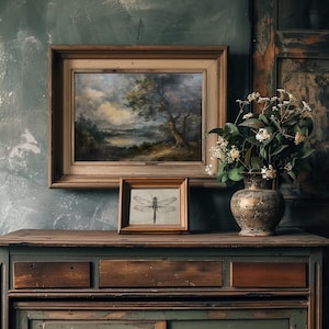 May include: A vintage wooden dresser with two drawers and a lower cabinet door. The dresser is painted a dark green colour and has a distressed finish. A framed landscape painting and a framed print of a dragonfly are displayed on top of the dresser. A vase with white flowers sits on the right side of the dresser.