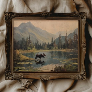 May include: An antique framed landscape painting depicts a moose standing in a lake with mountains and trees in the background. The painting has a vintage aesthetic with a brown ornate frame. The color palette includes blues, greens, and browns.
