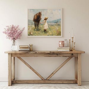 May include: A rustic wooden console table with a distressed finish. The table has a rectangular top and a unique X-shaped base. A framed painting of a girl and a horse in a field is hanging on the wall above the table. A vase with pink flowers, books, and decorative items are arranged on the table.