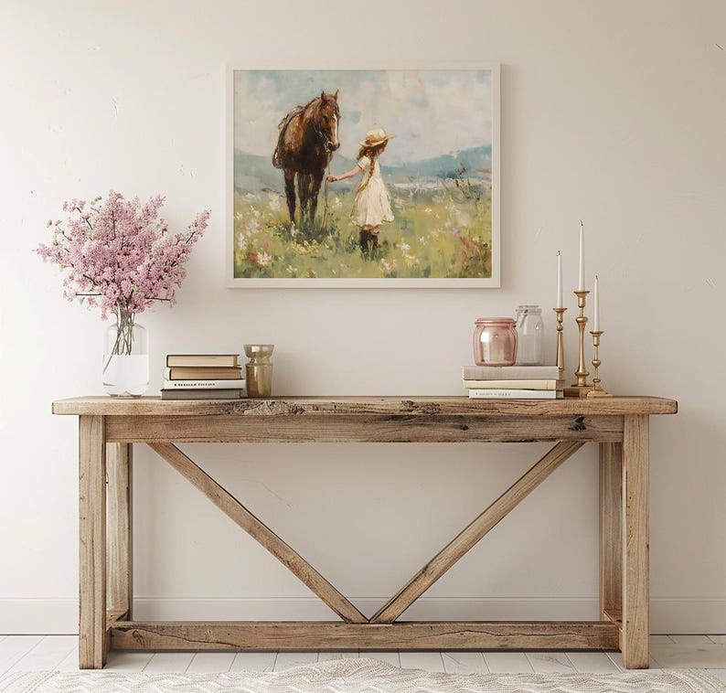 May include: A rustic wooden console table with a distressed finish. The table has a rectangular top and a unique X-shaped base. A framed painting of a girl and a horse in a field is hanging on the wall above the table. A vase with pink flowers, books, and decorative items are arranged on the table.