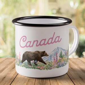May include: White enamel mug with a black rim and handle. The mug features the word "Canada" in pink script, a brown bear, a mountain, and colorful wildflowers. The mug is sitting on a wooden surface.
