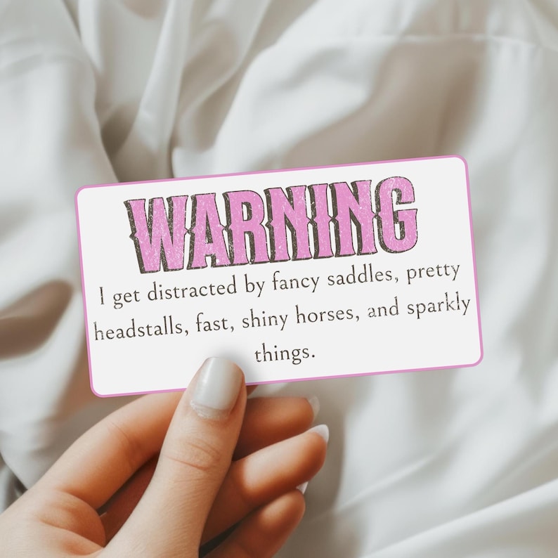 May include: A rectangular white sticker with a pink border featuring the text WARNING in large pink letters. Below, the text reads: I get distracted by fancy saddles, pretty headstalls, fast, shiny horses, and sparkly things. The sticker is held.