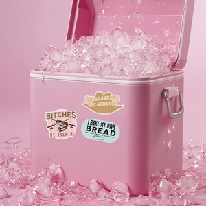 May include: A pink cooler with ice inside and three stickers on the side. The stickers say "Chase Sunsets Not Cowboys", "Bitches Be Fishin'" and "I Bake My Own Bread and I Don't Trust The Government".
