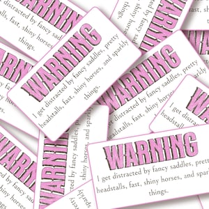 May include: A collection of rectangular white stickers featuring the word WARNING in bold pink letters. Below the text, each sticker reads: I get distracted by fancy saddles, pretty headstalls, fast, shiny horses, and sparkly things.