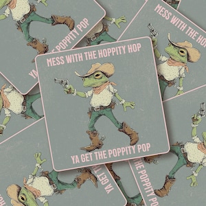 May include: A square sticker featuring a green frog dressed as a cowboy, wearing a tan hat, a white shirt with dark spots, and brown boots. The frog holds a silver revolver. The text reads: MESS WITH THE HOPPITY HOP YA GET THE POPPITY POP.
