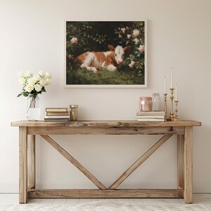 May include: A framed artwork depicts a brown and white calf resting in a lush green field surrounded by pink roses. The artwork is displayed above a wooden console table with decorative items.