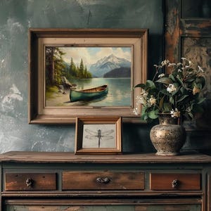 May include: A framed landscape painting of a lake with a green canoe, mountains, and trees. Below it, a smaller framed dragonfly sketch sits on a wooden dresser. A decorative vase with flowers completes the scene.