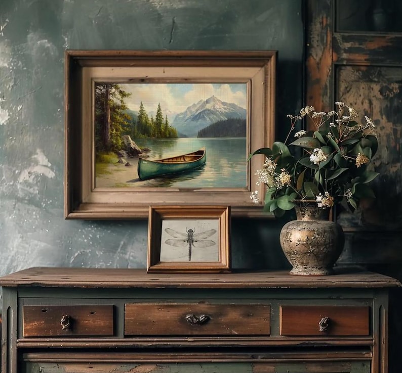 May include: A framed landscape painting of a lake with a green canoe, mountains, and trees. Below it, a smaller framed dragonfly sketch sits on a wooden dresser. A decorative vase with flowers completes the scene.