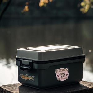 May include: A gray and black tackle box with a sticker that says "Bitches Be Fishin'" with a cartoon fish and fishing rod. The box is sitting on a wooden stump.