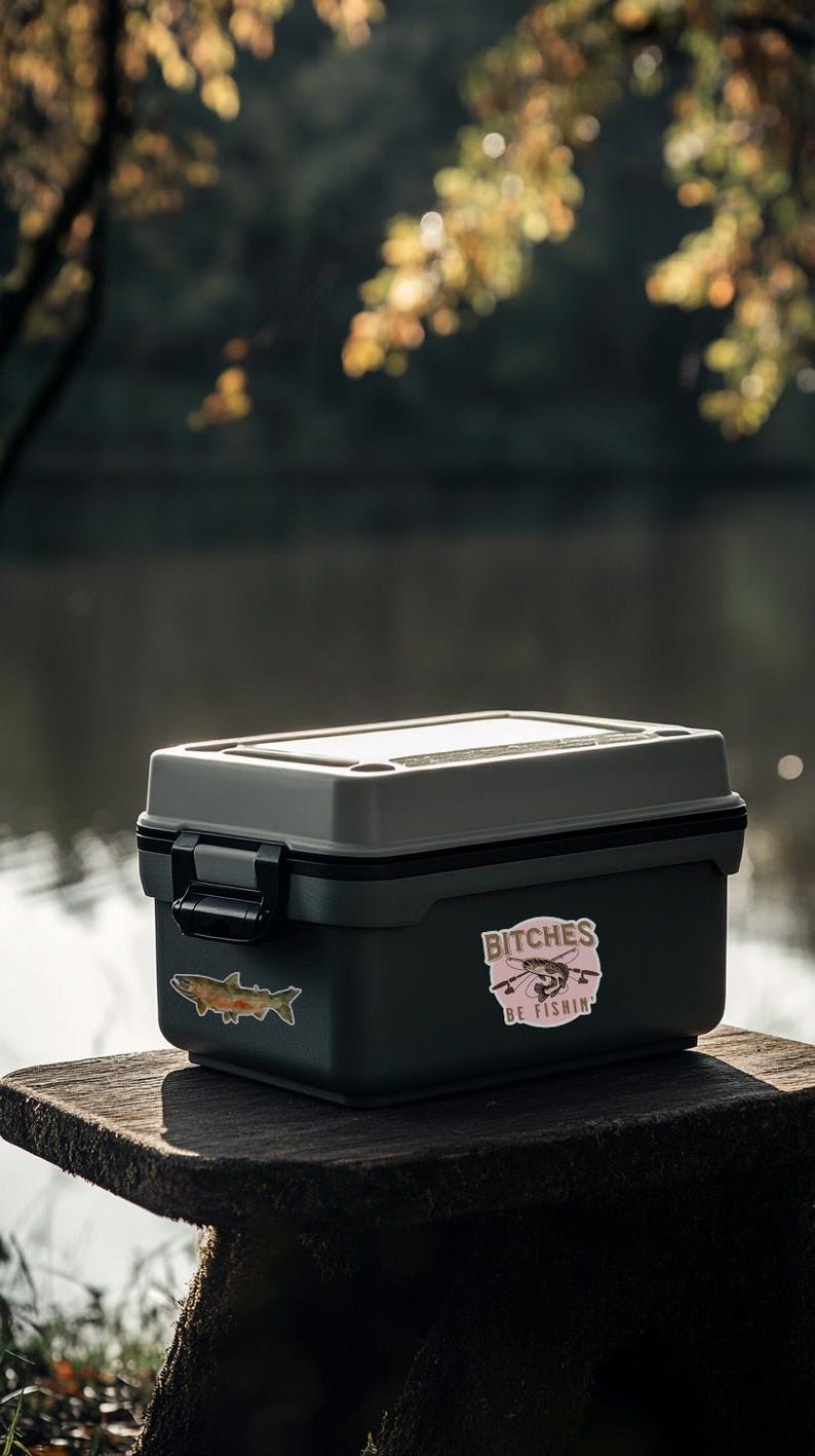 May include: A gray and black tackle box with a sticker that says "Bitches Be Fishin'" with a cartoon fish and fishing rod. The box is sitting on a wooden stump.