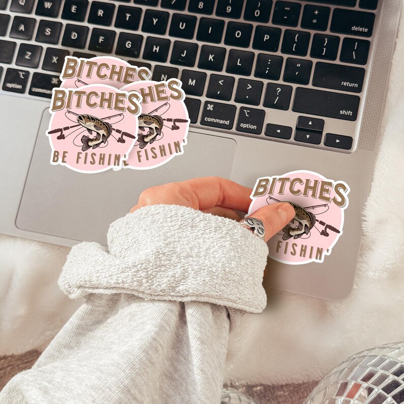 May include: Two pink and brown round stickers with the text "Bitches Be Fishin'" and a cartoon fish with fishing rods. A hand is holding a third sticker with the same design.