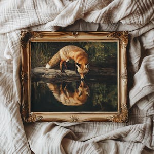 May include: A framed painting depicts a fox drinking from a body of water. The fox has orange fur and a white-tipped tail. The painting is in a gold ornate frame, resting on a neutral-colored fabric.