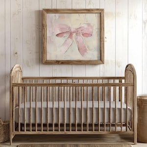 May include: A wooden crib with a gray linen mattress. The crib is in front of a white wood wall with a framed painting of a pink bow.