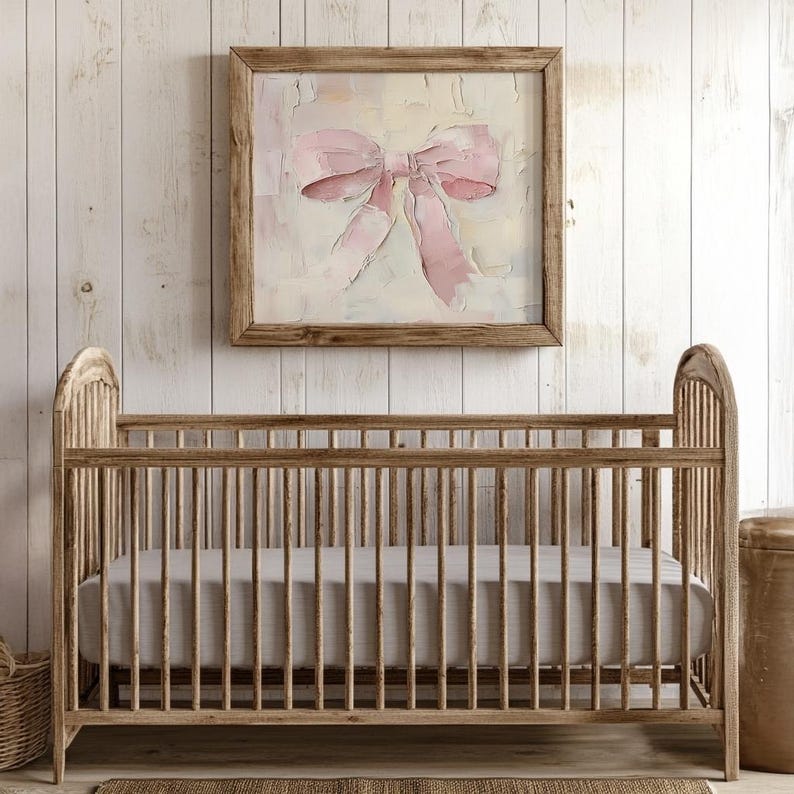 May include: A wooden crib with a gray linen mattress. The crib is in front of a white wood wall with a framed painting of a pink bow.
