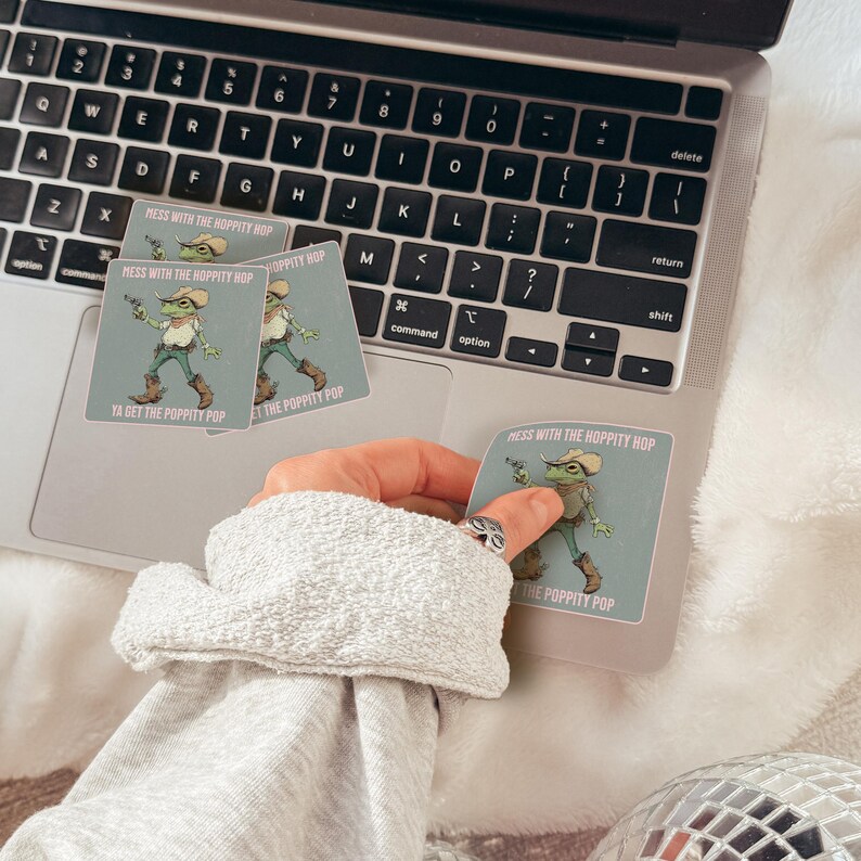 May include: Square stickers featuring a green frog dressed as a cowboy with a hat, vest, and boots, holding a small gun. The text reads: MESS WITH THE HOPPITY HOP YA GET THE POPPITY POP. The stickers are placed on the surface of a laptop keyboard.