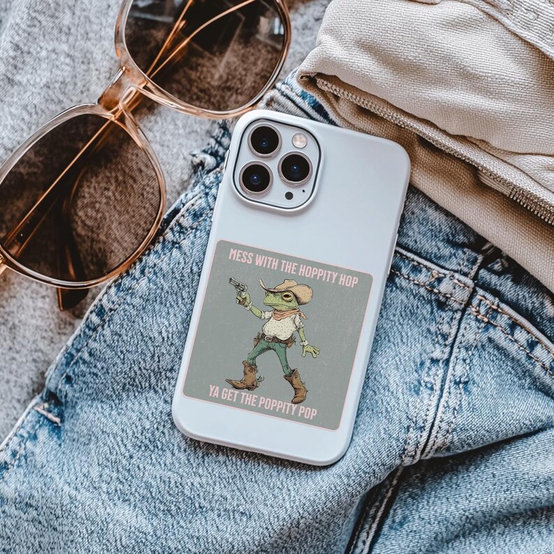 May include: A white smartphone case featuring a square sticker of a green frog dressed as a cowboy, wearing a hat, bandana, and boots, holding a pistol. The text reads: MESS WITH THE HOPPITY HOP YA GET THE POPPITY POP in a light pink font.