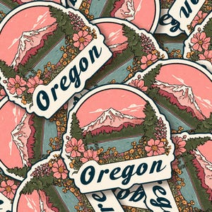 May include: A collection of Oregon-themed stickers. Each sticker features a mountain landscape with a pink sky, surrounded by flowers and trees. The word "Oregon" is written in a navy blue script.