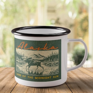 May include: White enamel mug with a black rim and handle. The mug features a green and tan graphic with the word "Alaska" in orange script, a moose, mountains, and the text "Land of the Midnight Sun."