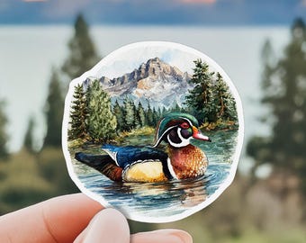May include: A circular sticker featuring a watercolor illustration of a wood duck swimming in a lake. The background displays a dense forest of green pine trees beneath a tall, rocky mountain peak under a soft, light-colored sky.