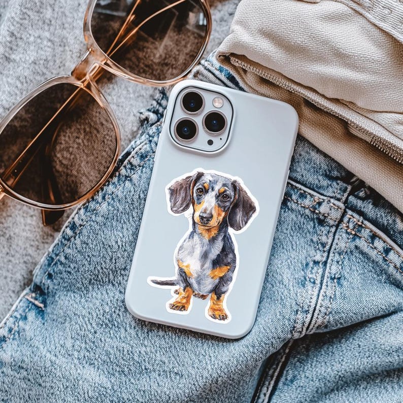 Puede incluir: Una funda para smartphone de color claro con una ilustraci&oacute;n detallada y colorida de un perro salchicha negro, fuego y blanco. El perro aparece en posici&oacute;n sentada con las orejas ca&iacute;das, mostrando texturas de pelaje y marcas realistas.