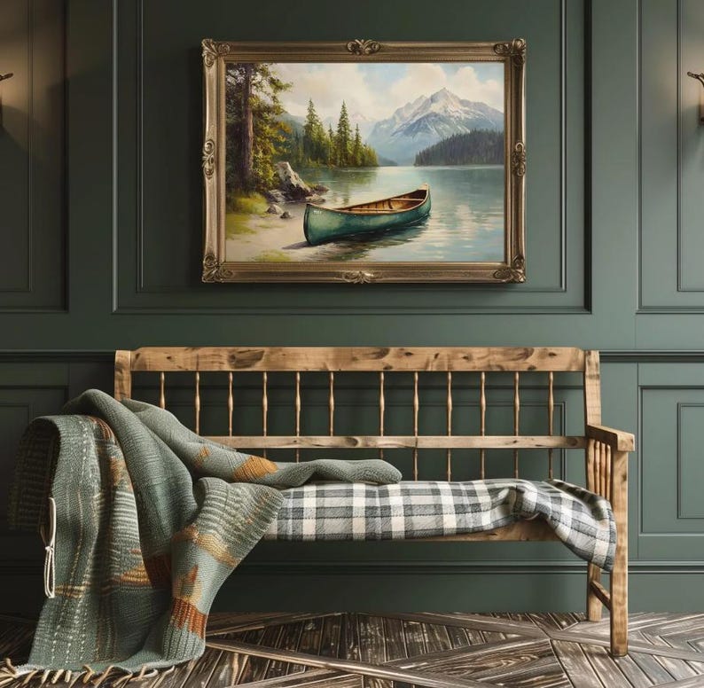 May include: A framed landscape painting depicts a teal canoe on a lake with mountains in the background. Below, a wooden bench with a plaid cushion is draped with a green and orange patterned blanket. The room has dark green walls and a wooden floor.