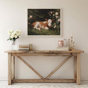 May include: A framed artwork depicts a brown and white calf resting in a lush green field surrounded by pink roses. The artwork is displayed above a wooden console table with decorative items.