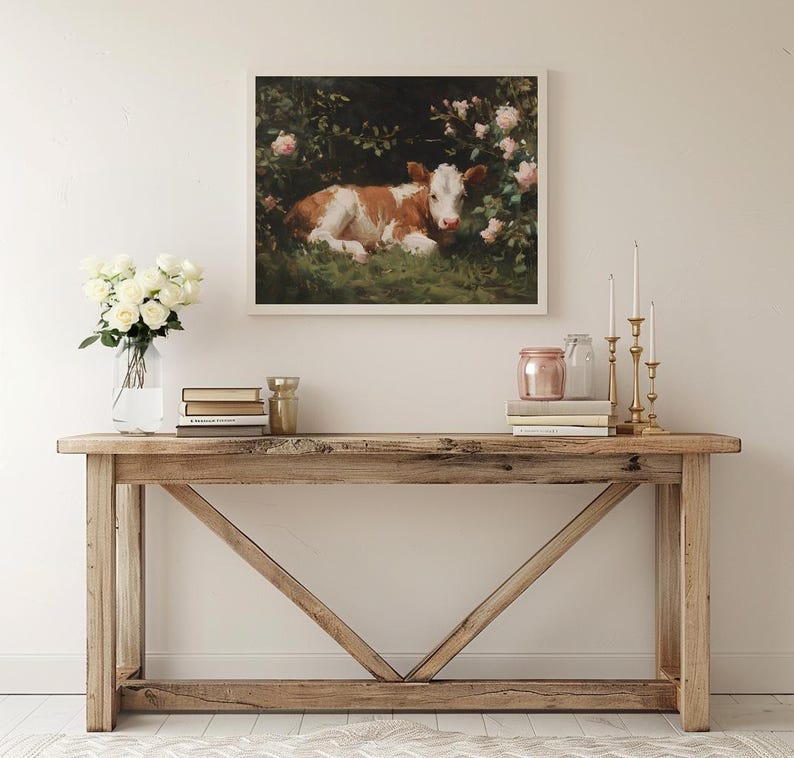 May include: A framed artwork depicts a brown and white calf resting in a lush green field surrounded by pink roses. The artwork is displayed above a wooden console table with decorative items.