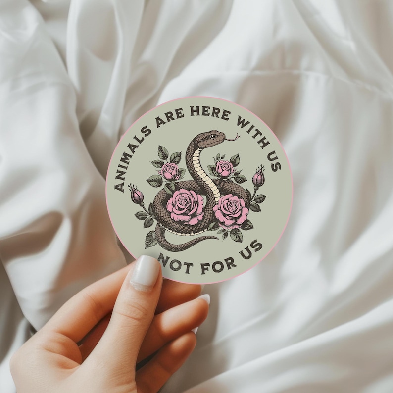 May include: Round sticker with a snake illustration surrounded by pink roses. The sticker has the text "ANIMALS ARE HERE WITH US NOT FOR US" in a circular pattern around the edge. The background is a light green color.