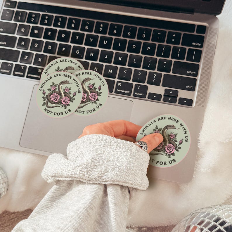 May include: Round stickers with a snake and rose design, and the text "ANIMALS ARE HERE WITH US NOT FOR US" in a circular layout. The stickers are light green with a black and pink color scheme. The stickers are on a laptop.