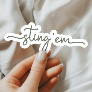 May include: A white sticker with the words "sting 'em" in a cursive font with a green outline. The sticker is held by a hand with a neutral background.