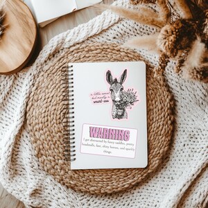 May include: A white spiral-bound notebook featuring two stickers. One sticker shows a donkey holding flowers with the text 'a little sweet but mostly a smart-ass'. The second sticker is a rectangular warning sign with pink text about horses.