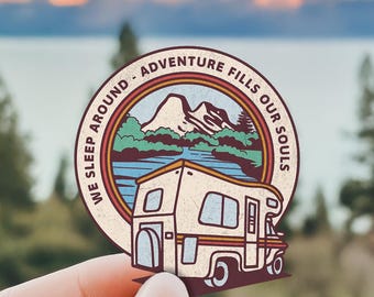 May include: A circular sticker featuring a cream-colored camper van in the foreground with a mountain and lake landscape in the background. The text WE SLEEP AROUND - ADVENTURE FILLS OUR SOULS is printed in a dark brown circular border.